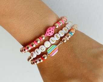 May include: Three gold bead bracelets with white letter beads spelling "PICKLEBALL" and a pink and white bead charm. The bracelets are stacked on a person's wrist.