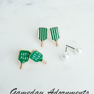 Puede incluir: Un conjunto de tres pares de pendientes. Dos pares presentan dijes en forma de paleta con rayas verdes y blancas con el texto "Let's Play". El tercer par presenta dijes de perlas blancas.