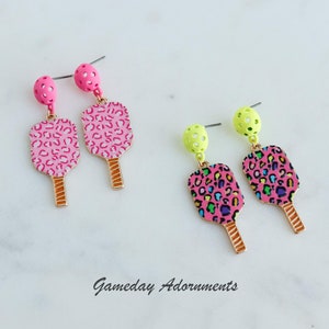 May include: A pair of dangle earrings featuring a pink and green pickleball paddle design with a leopard print. The earrings are made of metal and have a gold finish. The text "Gameday Adornments" is visible below the earrings.