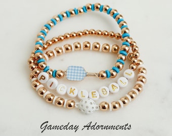 May include: A set of three gold beaded bracelets with a white and blue gingham pickleball paddle charm, white letter beads spelling "pickleball", and a white pickleball charm. The bracelets are stacked on a white marble surface. Gameday Adornments.