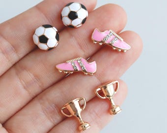 Puede incluir: Pendientes con temática deportiva, que incluyen balones de fútbol, botas de fútbol rosas con detalles de pedrería y tachuelas doradas con forma de trofeo. Los pendientes son pequeños y probablemente de metal con detalles de esmalte. El texto "Gameday Adornments" es visible.