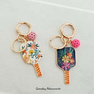 May include: Two gold-toned keychains featuring a floral patterned paddle design. One paddle is white with pink and yellow flowers, the other is navy blue with pink, yellow, and orange flowers. Each keychain has a small pink ball charm.