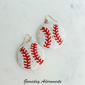 Puede incluir: Un par de pendientes de cuentas blancas con forma de pelotas de béisbol con costuras rojas. Los pendientes están sobre una superficie de mármol blanco con el texto "Gameday Adornments" debajo.