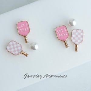 May include: Pink and white checked paddle and a pink paddle with the text "Let's Play" on it. The paddles are gold-toned and have a pearl-like ball next to them.