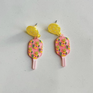 May include: A pair of pink and yellow earrings shaped like popsicles with a pineapple pattern. The pineapple has a smiling face with sunglasses and a heart.