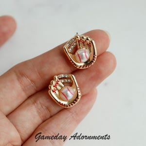 Könnte beinhalten: Goldene Baseballhandschuh-Ohrringe mit einem weißen Baseball und roten Nähten. Die Ohrringe befinden sich auf einer Hand.