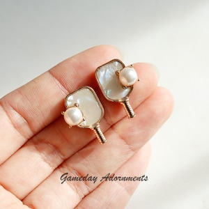 Puede incluir: Un par de pendientes de oro con forma de pala con un acento de perla. Los pendientes están hechos de madreperla blanca y tienen un acabado dorado.