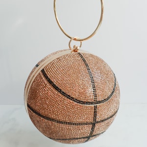May include: A basketball-shaped clutch with a gold handle. The clutch is covered in small, sparkling, copper-colored beads, with black lines mimicking a basketball's design. The text "Gameday Adornments" is at the bottom.