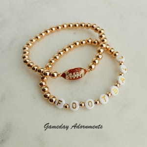 May include: Two gold beaded bracelets, one with a football charm and the other with white beads spelling out "FOOTBALL".