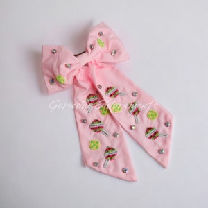 May include: A pale pink hair bow with a large bow and streamers. It features beaded lollipop and lime designs, plus scattered rhinestones. The text "Gameday Adornments" is printed on the bow. A decorative hair accessory.