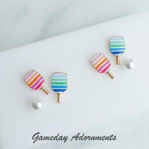 May include: A set of four colourful paddle earrings with rainbow stripes. The earrings are made of metal and have a gold finish. The earrings are displayed on a white surface with the text "Gameday Adornments".
