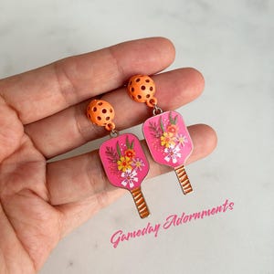 May include: A pair of pink and orange earrings featuring a floral design on a paddle-shaped charm. The earrings are hanging from a small orange ball with holes. The text "Gameday Adornments" is visible in the bottom right corner of the image.
