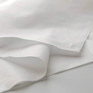 Könnte beinhalten: Nahaufnahme eines weißen, nicht gewebten Stoffes. Der Stoff ist weich und glatt, mit einer subtilen Textur. Er ist in der Mitte gefaltet, wodurch die Unterseite des Stoffes sichtbar wird.