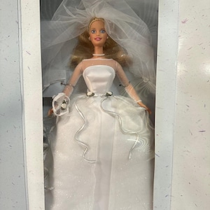 Puede incluir: Muñeca rubia con vestido de novia blanco y velo, expuesta en una caja. El vestido tiene escote corazón y detalles plateados. La muñeca lleva collar de perlas y tiara.
