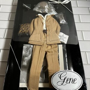 May include: A tan doll suit set including a blazer, trousers, and a white collared shirt, packaged in a black box with a Gene The Ashton-Drake Galleries oval label. The set includes a small leopard print accessory, a hat, and small metal items.