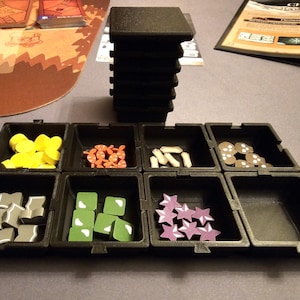 Könnte beinhalten: Ein schwarzer Plastik-Organizer mit acht Fächern, die jeweils verschiedene farbige Spielsteine enthalten. Der Organizer steht auf einer braunen Oberfläche.
