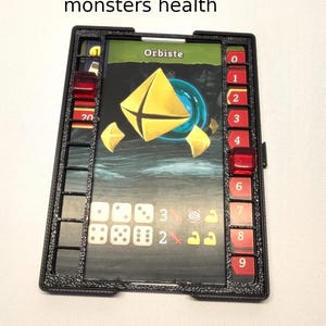 Puede incluir: Un rastreador de salud de pl&aacute;stico negro para un juego de mesa. El rastreador tiene una ranura para tarjetas, espacios para dados y una barra de salud numerada. La tarjeta muestra un orbe amarillo con el texto "Orbiste".