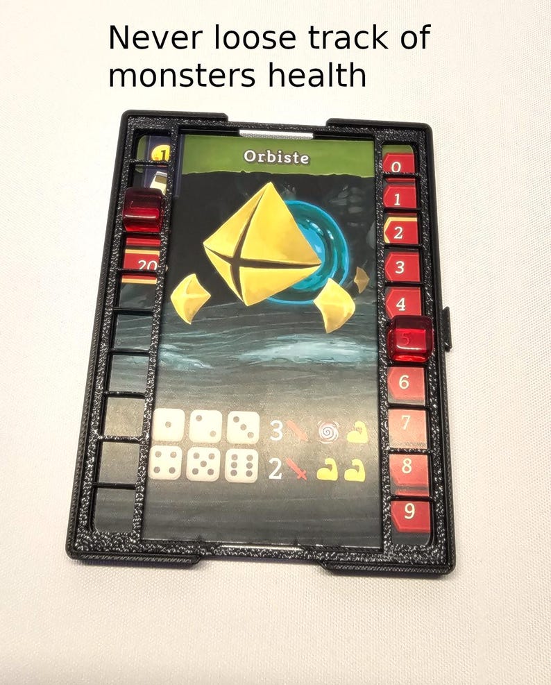 Puede incluir: Un rastreador de salud de pl&aacute;stico negro para un juego de mesa. El rastreador tiene una ranura para tarjetas, espacios para dados y una barra de salud numerada. La tarjeta muestra un orbe amarillo con el texto "Orbiste".
