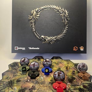 Op de afbeelding: De speldoos van The Elder Scrolls: La Trahison de la Deuxième Ère heeft een zwarte omslag met een metalen draken-ouroboros-logo. Daaronder toont een zeshoekig spelbord diverse karakterfiches in groene, blauwe, rode en bruine plastic houders.