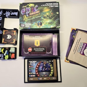 Puede incluir: Juego de mesa Tiny Epic Dungeons Adventures Phantom Voyage que incluye una caja de juego, organizadores de plástico negro, figuras en miniatura grises, un mazo de cartas con el texto The Phantom Voyage, una carta de personaje y una carta Tycoon Games.