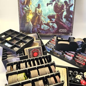 May include: A black plastic organiser with multiple compartments for storing game pieces and cards. The organiser is filled with various game components, including tokens, dice, and cards. The game box for Folklore: The Affliction is visible in the background.