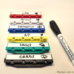 Op de afbeelding: Zes kleurrijke, gelabelde plastic containers met nummers op elk. De containers zijn verticaal gestapeld en gelabeld met "Life", "Armor", "Perception", "AP", "Skills" en "Cards". Een whiteboardmarker bevindt zich naast de containers.