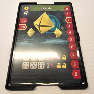 Puede incluir: Un porta tarjetas de pl&aacute;stico negro con una ventana de pl&aacute;stico transparente para mostrar una tarjeta de juego. La tarjeta es para un juego llamado "Orbiste" y presenta una forma geom&eacute;trica dorada sobre un fondo azul. El porta tarjetas tiene una escala num&eacute;rica del 0 al 9 en el lado derecho.