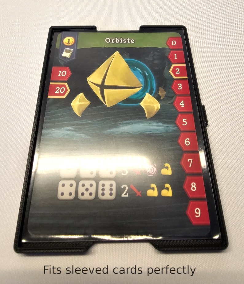 Puede incluir: Un porta tarjetas de pl&aacute;stico negro con una ventana de pl&aacute;stico transparente para mostrar una tarjeta de juego. La tarjeta es para un juego llamado "Orbiste" y presenta una forma geom&eacute;trica dorada sobre un fondo azul. El porta tarjetas tiene una escala num&eacute;rica del 0 al 9 en el lado derecho.