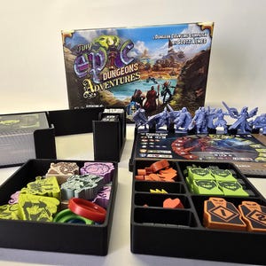 Op de afbeelding: Een bordspelset getiteld Tiny Epic Dungeons Adventures met een speldoos, zwarte plastic organizers, blauwe miniatuurfiguren, een spelbord en diverse gekleurde fiches, waaronder groene, oranje en rode vormen met gedrukte cijfers.