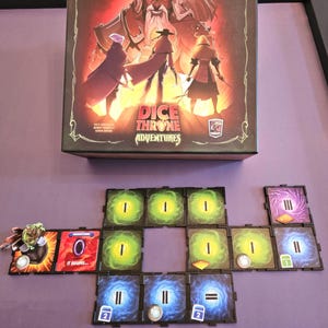 May include: A Dice Throne Adventures board game with a colorful box featuring four characters. The game board is laid out with various tiles, including green, blue, and purple tiles with symbols and numbers.