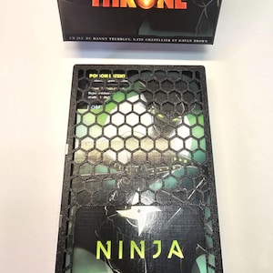May include: A board game box titled "DICE THRONE" in orange with a flame graphic. Below is a black game component with a hexagonal pattern and the word "NINJA" in yellow. The game components are black and green.