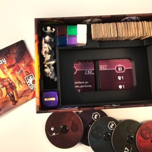 May include: A black plastic organizer insert for a board game box. The insert has compartments for cards, dice, and small game pieces. The insert is designed to keep the game components organized and easy to find. The insert is shown with a purple die, a stack of cards, and a few small plastic figures.