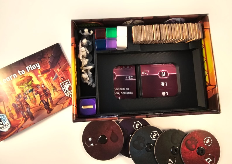 May include: A black plastic organizer insert for a board game box. The insert has compartments for cards, dice, and small game pieces. The insert is designed to keep the game components organized and easy to find. The insert is shown with a purple die, a stack of cards, and a few small plastic figures.