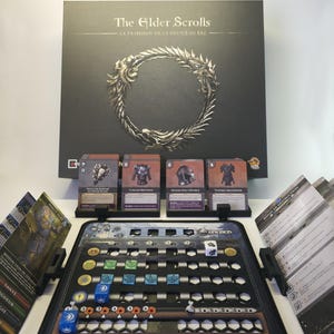 May include: A board game set, featuring a black box with the title "The Elder Scrolls" and a circular emblem. The set includes cards, game pieces, and a game board with various slots and markers. The game board is black and gray.