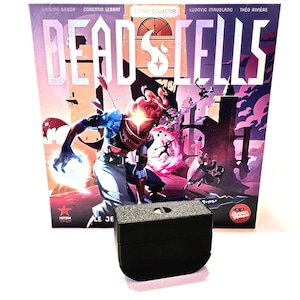 May include: A black plastic game stand with a textured surface. The stand is in front of a colorful game box with the title "Dead Cells" and the words "Edition Collector".