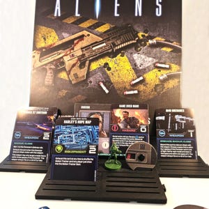 May include: A close-up of a board game set up for play. The game is based on the movie Aliens. The game board is made of black plastic with a yellow and black striped pattern. There are several cards on the board, including a weapon card, an equipment card, and a hazard card. There is also a miniature figure of a character from the movie. The game is set up for a player to take their turn.