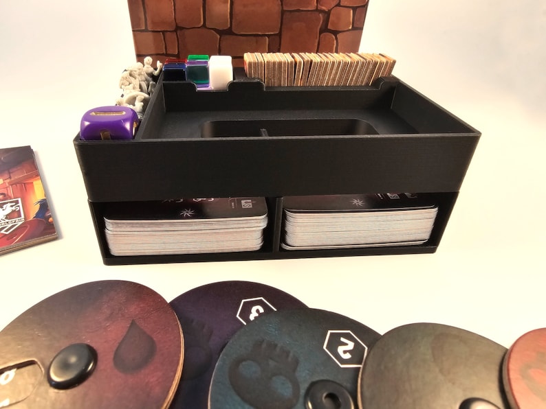 May include: A black plastic organizer with two compartments for storing cards, a top compartment for dice and tokens, and a small compartment for miniatures. The organizer is sitting on a white surface with several game tokens in the foreground.