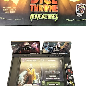 May include: A board game setup with the "Dice Throne Adventures" box and game components. The game includes character cards, dice, and a black plastic game organizer. The Blackroot Elf character card is visible, along with dice and health/combat point trackers.