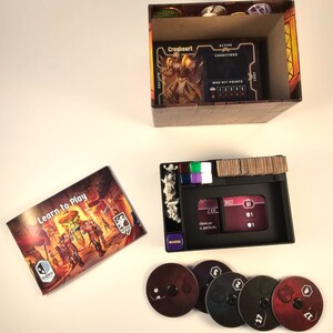May include: A board game with a learn to play guide, a box with a character card, a game board with tokens, and five black and red discs with numbers on them.