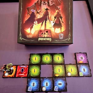 Può includere: Una scatola del gioco da tavolo Dice Throne Adventures con un'illustrazione rossa e nera di tre figure, posizionata sopra una disposizione di tessere quadrate con grafiche di portali verdi, blu e viola e piccoli gettoni.
