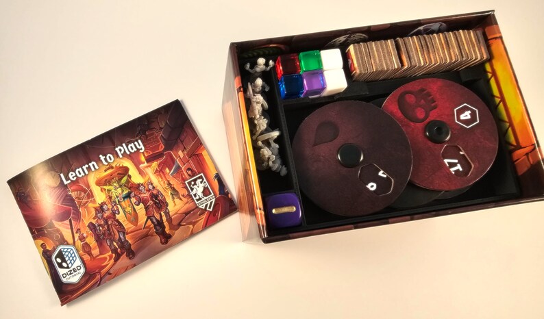 May include: A board game box with a brown and orange exterior. The box is open and shows the game's components, including dice, cards, and miniature figures. The box also includes a booklet titled "Learn to Play".