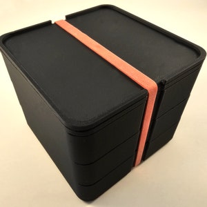 Peut inclure: Une boîte de rangement empilable noire avec un élastique rose. La boîte est en plastique et a une finition lisse et mate.