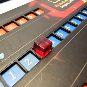 May include: A close-up of a red game piece on a black game board with numbered spaces. The numbers on the board are 1, 2, 3, 4, 5, 6, 7, 8, 9, and 0. The game piece is in the space marked 2.