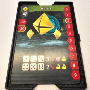 Puede incluir: Un porta tarjetas negro con una tarjeta de juego en su interior. La tarjeta tiene una forma geom&eacute;trica dorada con una cruz negra en el centro. La tarjeta tambi&eacute;n tiene un n&uacute;mero 1 en la esquina superior derecha y la palabra "Orbiste" en el centro superior. El porta tarjetas tiene una escala num&eacute;rica en el lado derecho, del 0 al 9.