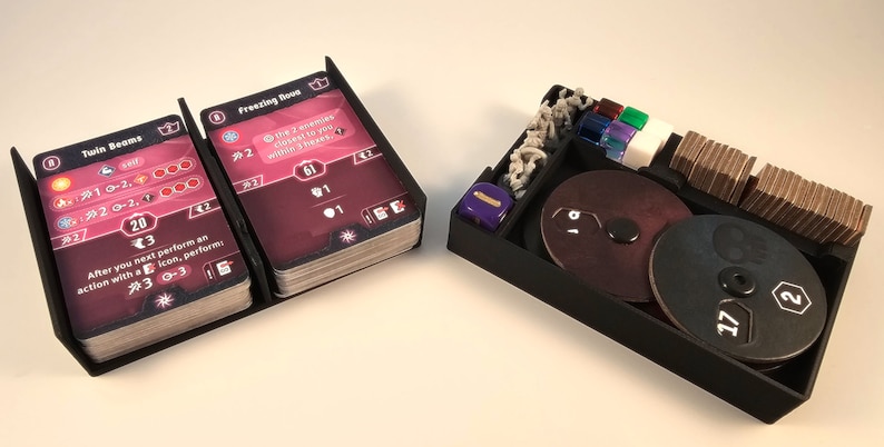 May include: A black plastic organizer with two card holders, two circular trays, a dice tray, and a stack of cards. The card holders have the text "Twin Beams" and "Freezing Boost" on them. The circular trays have the numbers 17, 18, and 19 on them. The dice tray has a purple die in it.