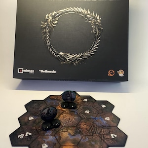 May include: A board game box with the title "The Elder Scrolls" and the text "LA TRAHISON DE LA DEUXIÈME ÈRE". The box is black with a silver emblem. A hexagonal game board and game pieces are in front of the box.