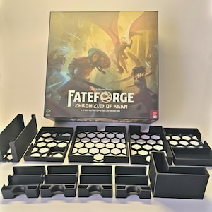 May include: A board game box titled "Fateforge: Chronicles of Kaan" with fantasy artwork. Black plastic game organisers are arranged in front of the box. The box art features figures in a battle scene with a dragon.