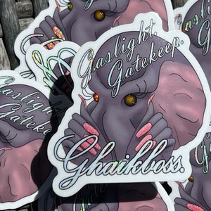 Peut inclure: Un autocollant avec un éléphant de dessin animé à la peau rose et violette, de longs cils et du vernis à ongles rose. L'éléphant se couvre la bouche avec ses mains. Le texte "Gaslight Gatekeep Girlboss" est écrit en cursive blanche sur l'autocollant.
