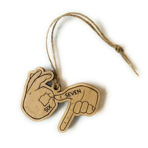 May include: Wooden ornament depicting the number seven in American Sign Language. The ornament is light brown with black outlines and the words "SIX" and "SEVEN" are printed on it. A natural twine loop is attached for hanging.