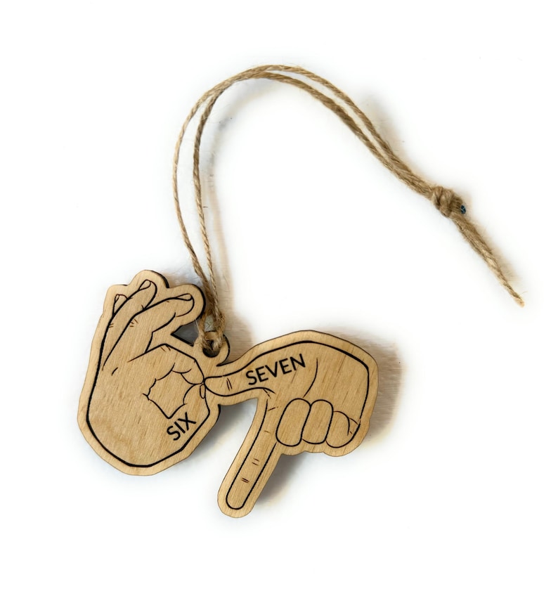 May include: Wooden ornament depicting the number seven in American Sign Language. The ornament is light brown with black outlines and the words "SIX" and "SEVEN" are printed on it. A natural twine loop is attached for hanging.
